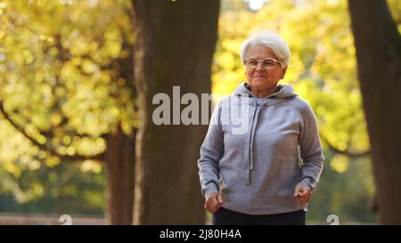 Jogging nel parco. Signora caucasica in pensione dai capelli grigi in felpa con cappuccio grigia corre intorno al suo parco preferito. Concetto di benessere. Foto di alta qualità Foto Stock