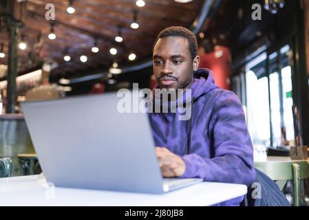 Freelancer maschile africano americano concentrato indossando felpa con cappuccio casual seduto al tavolo e digitando su netbook mentre si lavora su un progetto online in caffetteria Foto Stock