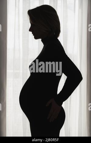 Vista laterale della silhouette giovane donna incinta in piedi contro la finestra a casa. Ritratto in attesa di donna in camera oscura. Foto Stock