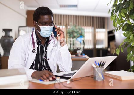Medico afroamericano in maschera che parla sul telefono cellulare Foto Stock