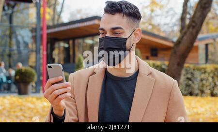 Giovane spagnolo in maschera medica parlare in videochiamata utilizzando smartphone in piedi nel parco ragazzo ispanico annuisce con approvazione guardando lo schermo del telefono Foto Stock