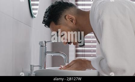 Vista laterale arab spaniard indiano uomo arabo ragazzo apre rubinetto vicino lavandino a casa in bagno dell'hotel lavandes faccia con acqua calda fredda mattina bellezza Foto Stock