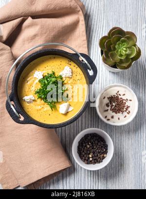 Una zuppa di purea di zucca in un recipiente in ghisa con pezzi di formaggio bianco e prezzemolo tritato sulla parte superiore di un tavolo grigio in legno. Panno di tovagliolo beige, crema bianca Foto Stock