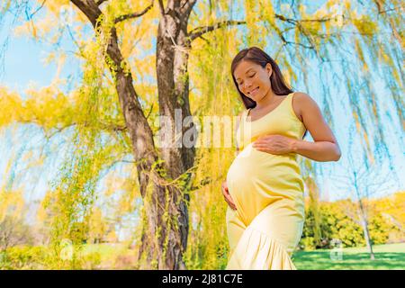 Donna incinta asiatica sorridente guardando giù il suo bump del bambino per la gravidanza di maternità photoshoot in giallo parco di primavera natura all'aperto. Sano felice Foto Stock