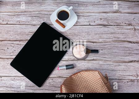 Tablet sul tavolo circondato da una tazza di caffè, un rossetto e una borsa per il trucco vista dall'alto. Foto Stock