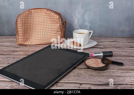 Tavoletta sul tavolo circondata da rossetto, polvere pressata, tazza di caffè e borsa per il trucco vista laterale. Foto Stock