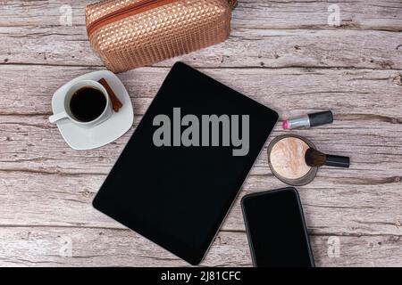 Tablet sul tavolo circondato da smartphone, rossetto, polvere pressata, tazza di caffè e borsa per il trucco vista dall'alto. Foto Stock