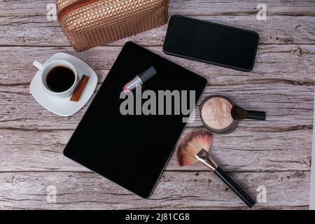 Rossetto sul tablet circondato da una tazza di caffè, una borsa per il trucco, uno smartphone, una polvere premente e una vista dall'alto con spazzola. Foto Stock