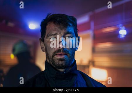 Primo piano di un pompiere sporco con un camion dei pompieri in background di notte. Foto Stock
