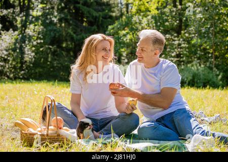 Coppia caucasica gioiosa siede nel parco cittadino. Uomo e donna all'aperto, abbracciante, sorridente e ridente in una giornata estiva soleggiata in abiti casual leggeri. Felice coppia pensionato concetto Foto Stock