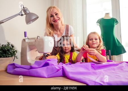 Sorridendo felice madre bionda multitasking insegna a piccole sorelle a cucire su una macchina da cucire da sola Foto Stock