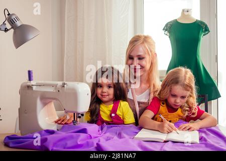 Sorridendo felice madre bionda multitasking insegna a piccole sorelle a cucire su una macchina da cucire da sola Foto Stock