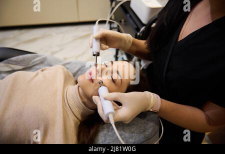 Primo piano una graziosa donna caucasica di mezza età riceve un trattamento di microcorrente facciale dall'estetista al salone termale. Cosmetologo che utilizza impu elettrico Foto Stock