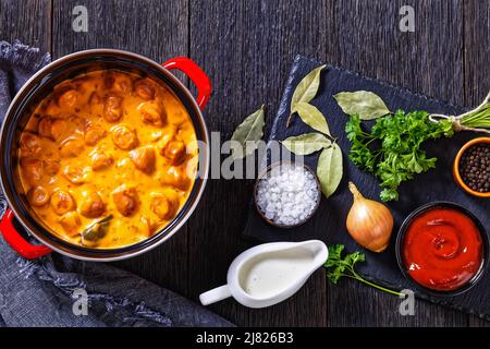 nakkikastike, salsicce finlandesi, amalgia in salsa di pomodoro cremosa in pentola rossa su tavola di legno scuro con ingredienti, vista orizzontale dall'alto, posa piatta, clo Foto Stock