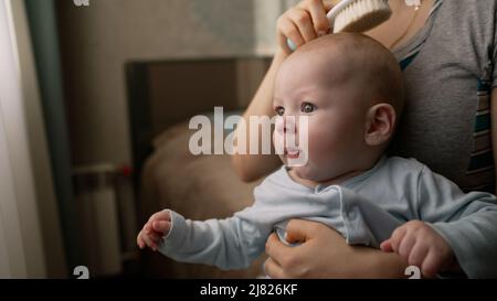 Mamma combatte il bambino tra le braccia. Una madre pettina i capelli del ragazzino. Un ragazzo carino è seduto sul grembo di sua madre. Madre con bambino. Foto Stock