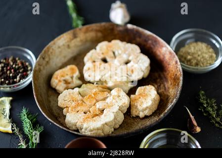 Cucina con bistecca di cavolfiore. Il cavolfiore crudo cosparso di spezie si trova in una padella. Olio d'oliva, erbe, varie spezie nelle vicinanze. Sfondo scuro. Foto Stock