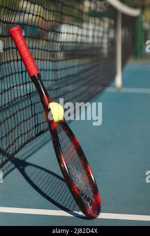 Palla con racket rosso appoggiato su rete da tennis in campo durante la giornata di sole Foto Stock