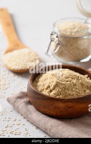 Farina di sesamo in ciotola di legno e semi in vaso di vetro su sfondo bianco. Verticale. Primo piano. Foto Stock