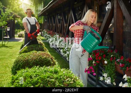 Bella donna caucasica con capelli biondi che annaffiano fiori in pentole sul cortile posteriore. Uomo robusto e bello in occhiali e guanti di sicurezza con trimmer elettrico per la sagomatura dei cespugli. Lavori stagionali in giardino. Foto Stock