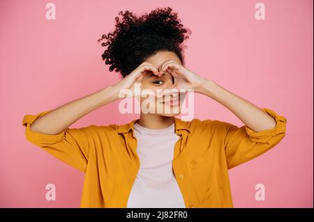 Bella candida ragazza afro-americana piacevole, fa il cuore gesto con le mani vicino occhio, dimostra segno d'amore, si leva su isolato sfondo rosa, guardando la macchina fotografica anche se gesto d'amore, sorriso amichevole Foto Stock