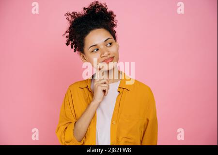 La ragazza curly positiva pensierosa afroamericana abbastanza haired in un vestiti casual, pensando ad un'idea, guardando via pensivamente con il dito vicino alla bocca, progettando, levandosi in piedi su sfondo rosa isolato Foto Stock