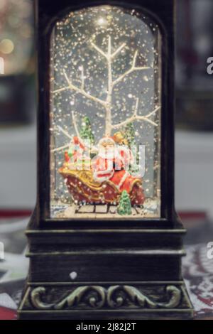 Lanterna di Natale in stile retrò con la statuetta di santa su una slitta e la neve cadente dai luccicanti canfetti. Immagine sfocata. Foto Stock