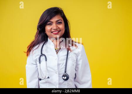 Donna indiana giovane e bella donna bionda medico ginecologo utilizzando stetoscopio in un cappotto medico bianco su sfondo giallo in studio Foto Stock