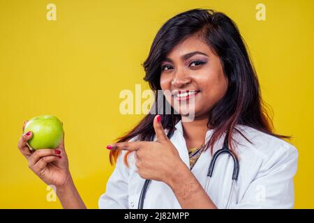 Donna indiana giovane e bella bionda medico ginecologo utilizzando stetoscopio tenendo una mela verde in mano in un cappotto medico bianco su un giallo Foto Stock