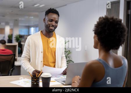Sorridente uomo d'affari afroamericano con documenti e laptop in cerca di donna d'affari sul posto di lavoro Foto Stock