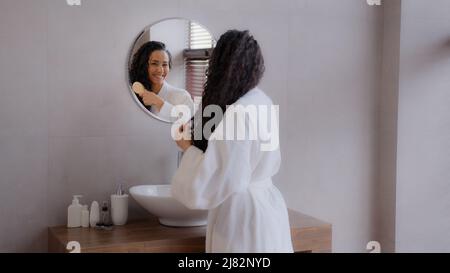 Giovane donna felice in piedi in bagno guardando in specchio toccare il viso dopo l'applicazione di crema gode di pelle sana idratata ammirando se stessa danza Foto Stock