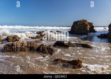 Spiaggia rocciosa a Malibu, California. Onde lavarsi verso la riva. Acqua, schiuma e alghe in primo piano. Cielo blu e Oceano Pacifico in dista Foto Stock