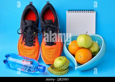 Sneakers arancioni, nastro graduato, bottiglia d'acqua, blocco note e ciotola di frutta a forma di cuore su sfondo blu. Programma di dimagrimento Foto Stock