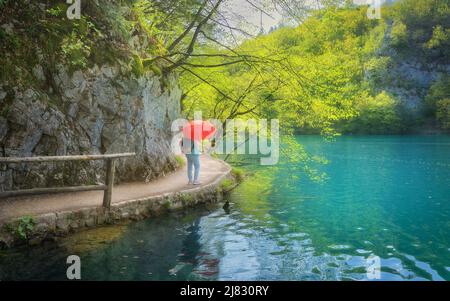 Donna con ombrello rosso sul sentiero vicino al bel lago in pioggia Foto Stock