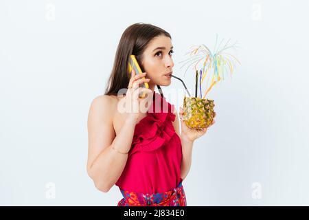 Brunette ragazza alla moda drink ananas cocktail con cannuccia e parla sul telefono cellulare mentre in piedi su sfondo bianco Foto Stock