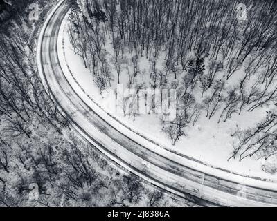 Vola sopra la curva invernale della neve. Strada bianca e nera circondata da foresta coperta di neve. Vista aerea verso il basso Foto Stock