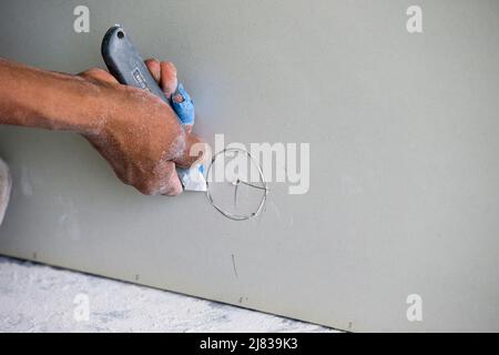 Il lavoratore di costruzione / gruccia di Drywall sta tagliando e segando un intero nella parete asciutta - pietra di foglio per l'installazione intorno ad un ostacolo Foto Stock