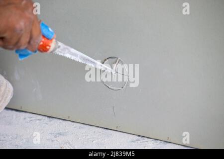 Il lavoratore di costruzione / gruccia di Drywall sta tagliando e segando un intero nella parete asciutta - pietra di foglio per l'installazione intorno ad un ostacolo Foto Stock