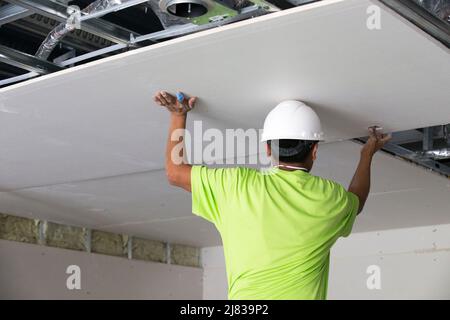Installer di drywall mettendo e fissando un grande foglio di drywall sul soffitto di un edificio in costruzione Foto Stock