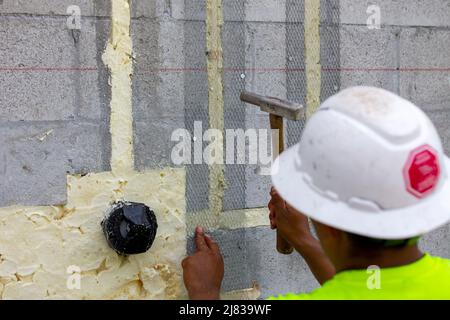 un operatore edile sta fissando una rete in acciaio per l'applicazione in stucco con un martello e tasselli in calcestruzzo, nascondendo tubi idraulici Foto Stock