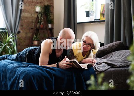 Moderna coppia matura trascorrere il tempo rilassandosi a letto a casa leggendo romanzo e discutendolo Foto Stock