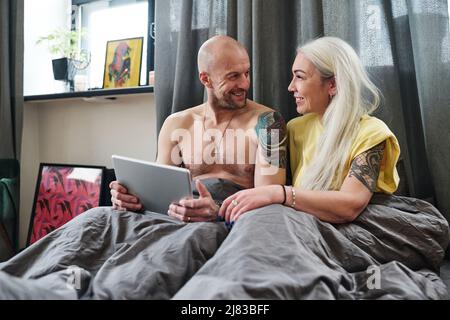 Gioioso tatuato uomo e donna maturi seduti a letto al mattino guardando qualcosa di divertente in Internet su tablet digitale Foto Stock