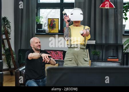 Uomo caucasico maturo seduto sul divano guardando sua moglie a giocare al videogioco utilizzando visore VR e controller Foto Stock