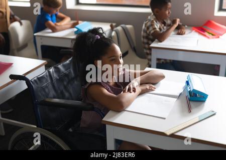 Sorridente studentessa biraciale seduta su sedia a rotelle alla scrivania in classe Foto Stock