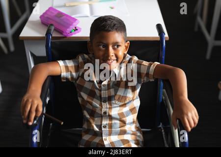 Vista ad alto angolo del ragazzo biraciale elementare sorridente seduto su sedia a rotelle in classe Foto Stock