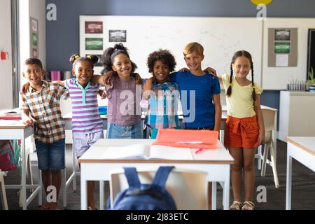 Ritratto di sorridenti studenti della scuola elementare multirazziale in piedi con il braccio intorno in classe Foto Stock