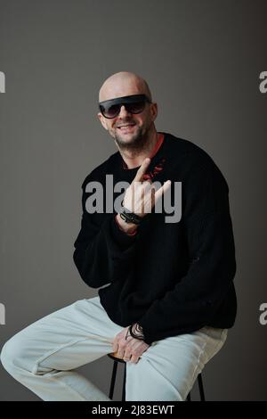 Ritratto verticale dello studio di allegro uomo caucasico seduto su sedia indossare occhiali da sole mostrando corna segno gesto sorridente alla macchina fotografica Foto Stock