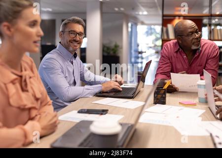 Ritratto di un uomo d'affari caucasico sorridente seduto tra colleghi durante l'incontro sul posto di lavoro Foto Stock
