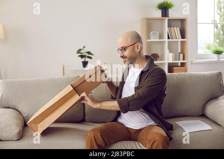 Uomo felice seduto sul divano a casa e disimballare il sacchetto di consegna della carta dal deposito in linea Foto Stock