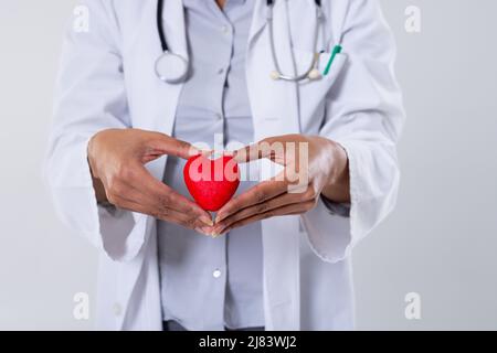 Metà della sezione di medico di metà adulta afroamericana che tiene il cuore rosso su sfondo bianco Foto Stock