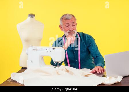 piacevole uomo vecchio che lavora in studio di sartoria facendo vestiti alla macchina da cucire, sfondo giallo Foto Stock